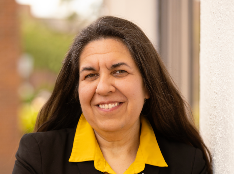 A woman wearing a collared yellow shirt and black blazer stands outside smiling at the camera.