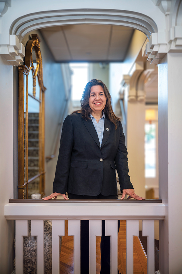 A woman in a business suit stands in a large hallway. There is a banister in front of her and a staircase behind her.