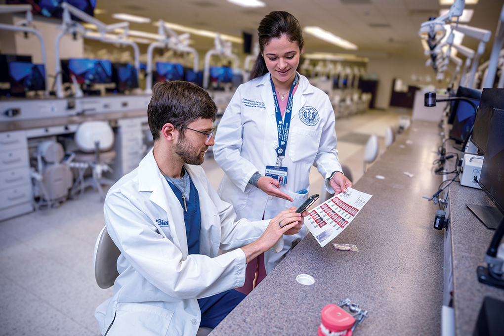 A female dentist wearing a lab coat stands beside a seated male dental student wearing a lab coat and holds up a sheet of paper with a QR code for the student to scan.