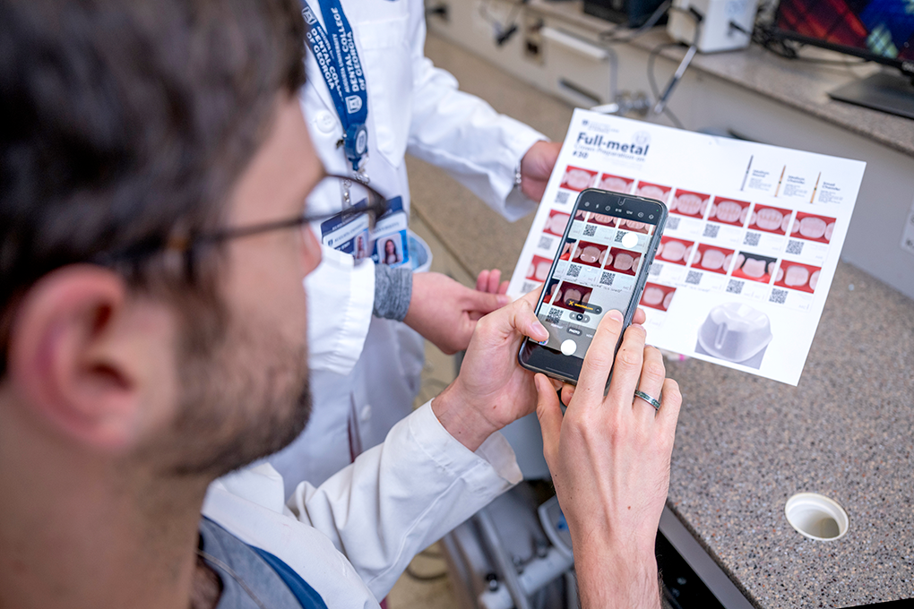 A male dental student wearing glasses and a lab coat scans a QR code on a crown preparation manual.