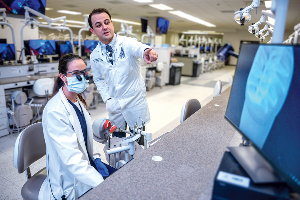 A male dentist wearing a lab coat stands behind a female dental student wearing a lab coat and points to a digital model of a tooth on a computer monitor.
