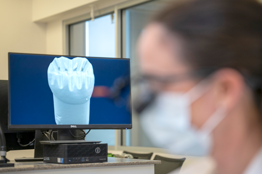 A computer monitor showing a 3D image of a molar sits behind an out-of-focus dental student.