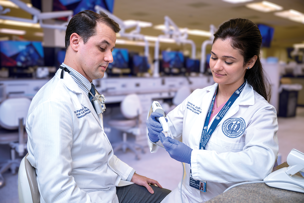 A male dentist wearing a lab coat and a female dentist wearing a lab coat use an intraoral scanner to create a 3D image of a plastic tooth.