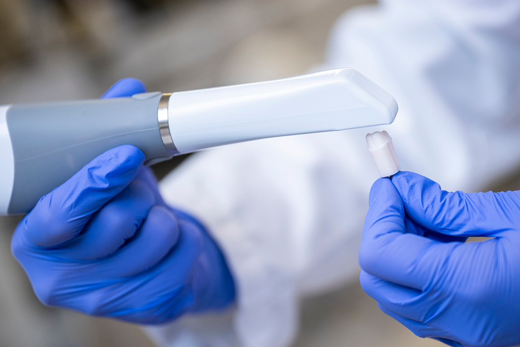Gloved hands hold an intraoral scanner and scan a plastic tooth for digital modeling.