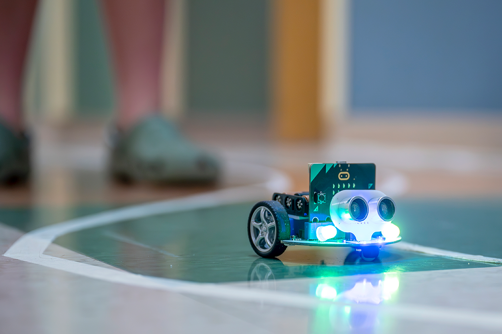 A mini robot car with headlights sits on the ground while two boys stand behind it trying to drive it.