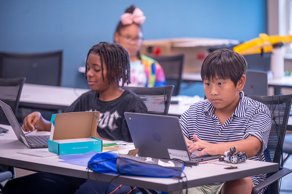 Two young boys sit at a desk and code software on laptops.