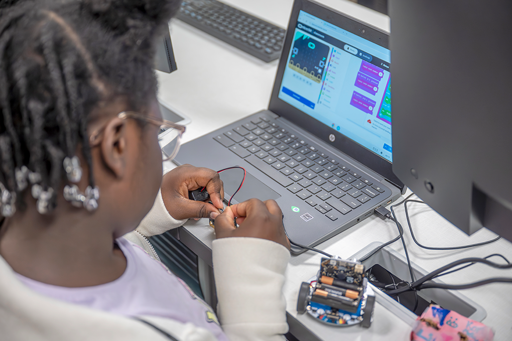 A young girl sits in front of a laptop and uses coding software to program a remote controlled miniature vehicle.