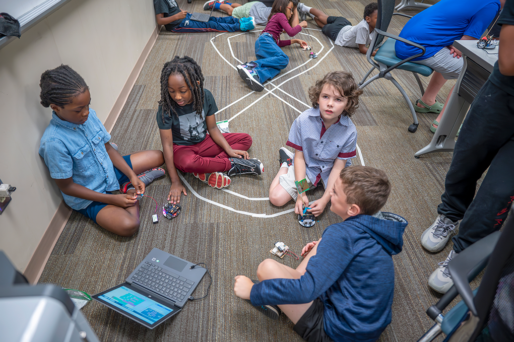 A group of young children sit on the floor in front of a race track made from tape and talk to each other.