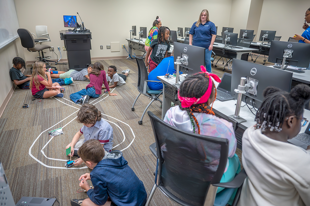 A group of kids sit in a classroom and practice coding and operating mini robot cars.