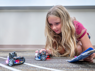 A young girl lies on the floor and examines two miniature robotic cars.