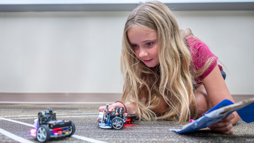 A young girl lies on the floor and examines two miniature robotic cars.