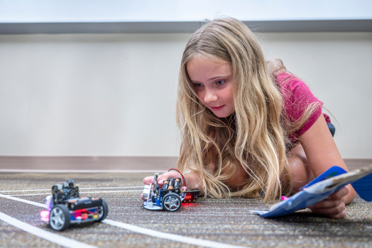 A young girl lies on the floor and examines two miniature robotic cars.
