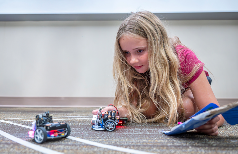 A young girl lies on the floor and examines two miniature robotic cars.