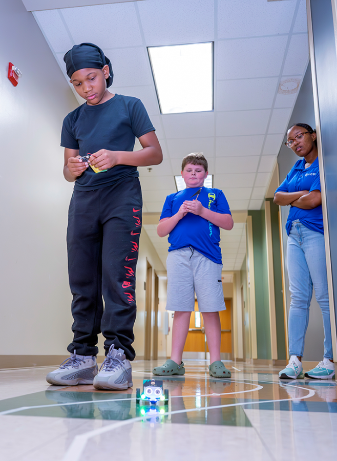 A young boy operates a miniature remote controlled car while another young boy and a female teacher stand behind him and watch.