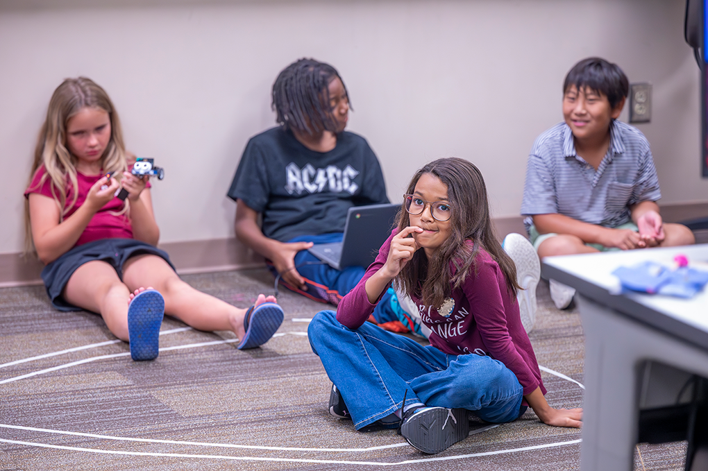 A group of young kids sit on the floor and listen to what their teacher is telling them.