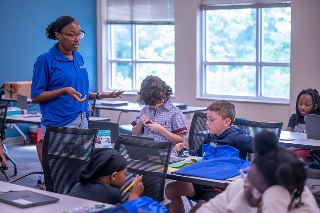 A female teacher stands and talks to kids sitting at desks in a classroom.