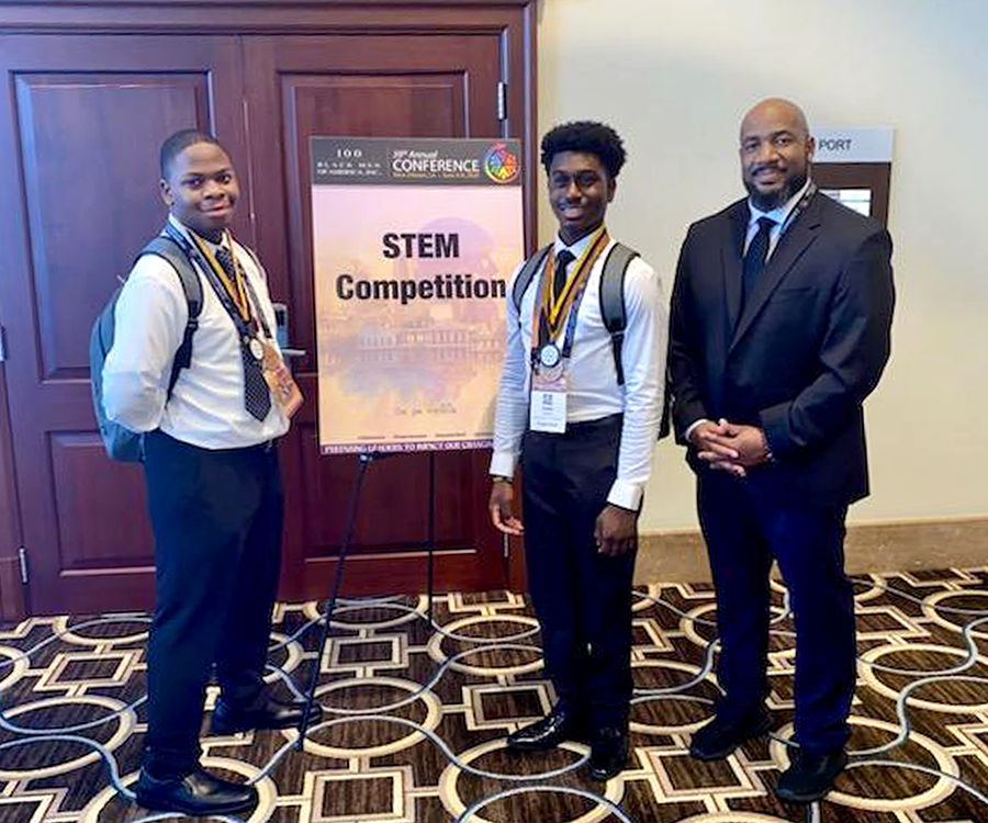 Two male high school students stand with an older male advisor outside of a conference room. There is a sign that reads "STEM Competition."