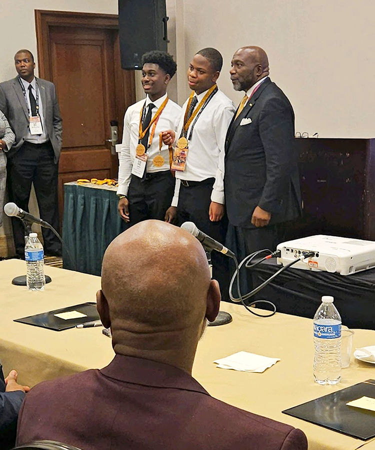 Two male high school students stand with an older man stand in front of a large full of people. The two students are holding medals for finishing third in a competition.