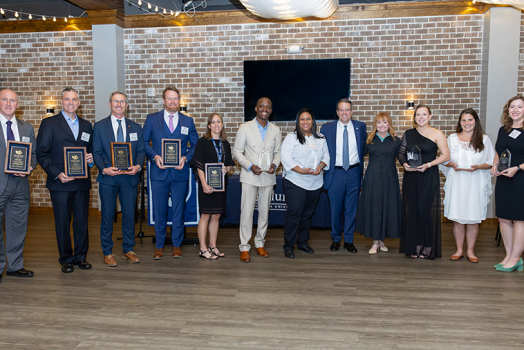 A group of people hold awards and in a large banquet room in front of a larger group of people.