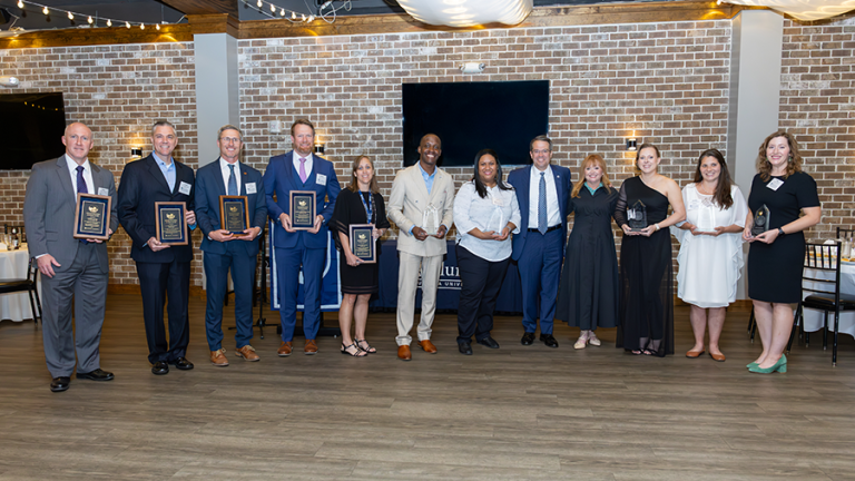 A group of people hold awards and in a large banquet room in front of a larger group of people.
