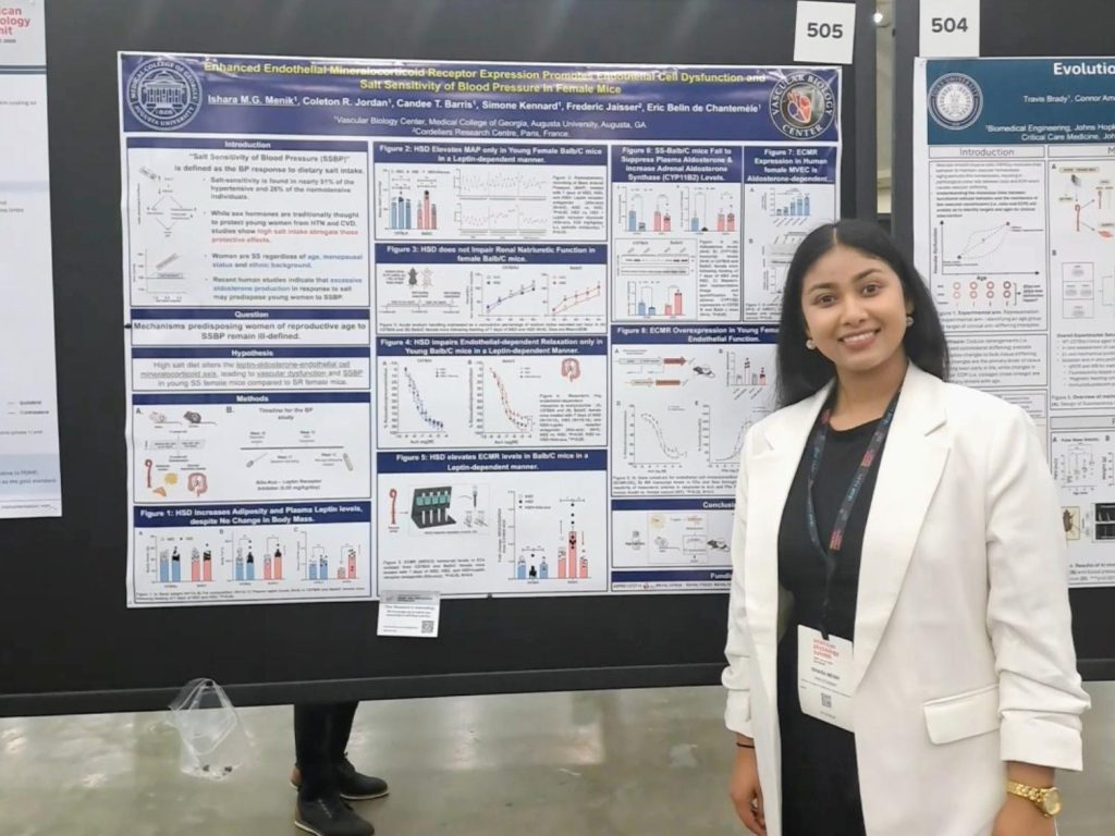 A woman in a dark dress and white jacket with a badge hanging from her neck stands in front of her research poster