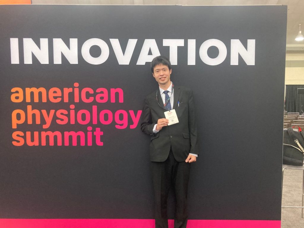 A man holds the badge hanging around his neck in his hand and stands in front of a sign reading "INNOVATION american physiology summit"