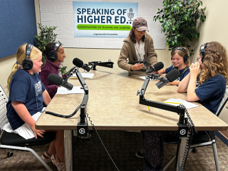 Four young girls sit around a table with large podcasting microphones. They are all wearing headphones to learn what it is like to host a podcast.