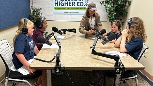 Four young girls sit around a table with large podcasting microphones. They are all wearing headphones to learn what it is like to host a podcast.