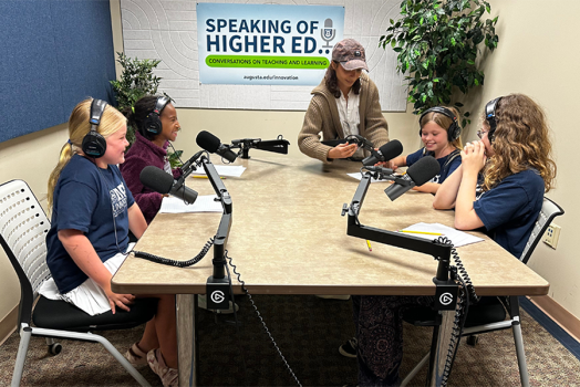 Four young girls sit around a table with large podcasting microphones. They are all wearing headphones to learn what it is like to host a podcast.