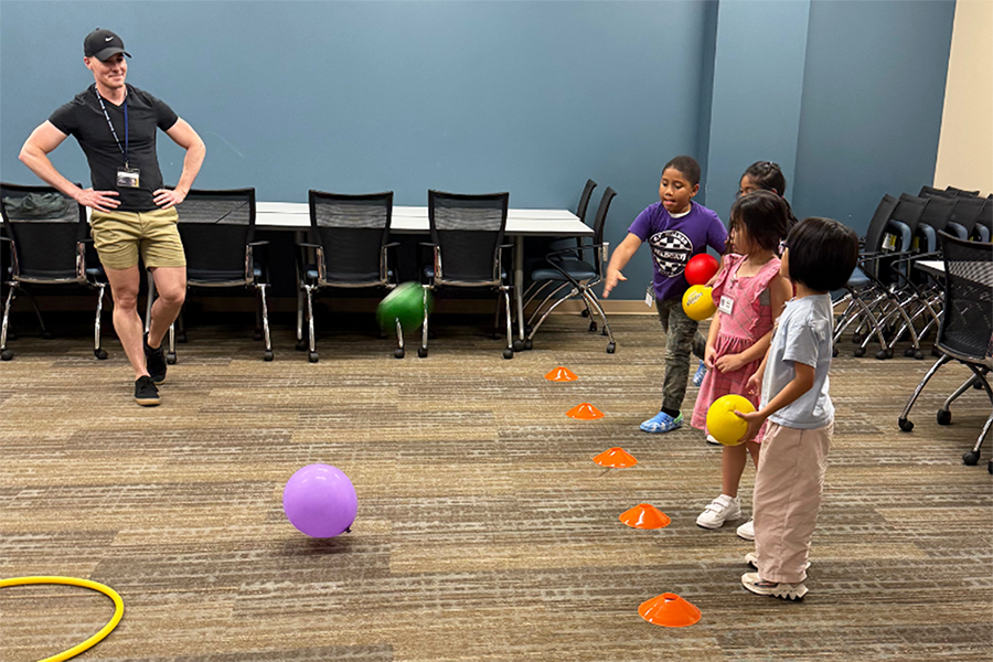 Four elementary-aged children play a game during a summer camp. They each have a couple balls and are throwing them at a balloon to move it into a hoop on the floor.