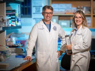 A male and female dentist in lab coats and glasses pose in their laboratory and smile at the camera.