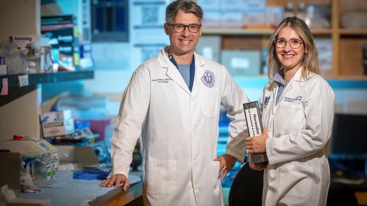 A male and female dentist in lab coats and glasses pose in their laboratory and smile at the camera.