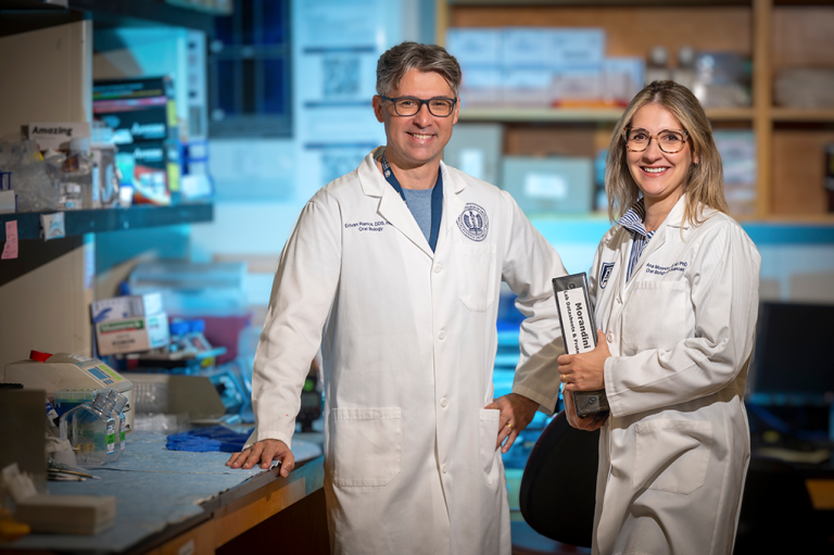 A male and female dentist in lab coats and glasses pose in their laboratory and smile at the camera.