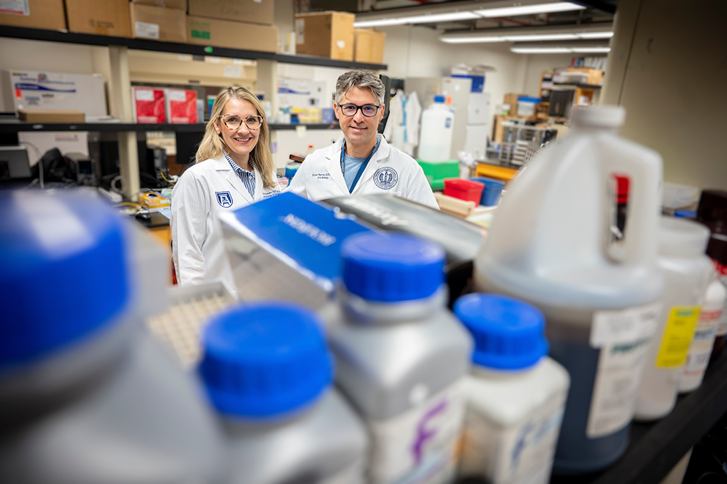 A male and female dentist wearing lab coats and glasses pose in their laboratory behind chemical bottles.