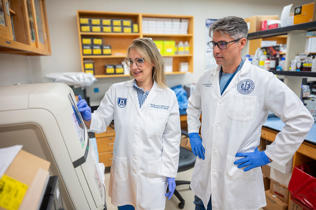 A male and female dentist wearing lab coats and glasses stand in a laboratory and examine a screen on an electronic machine.
