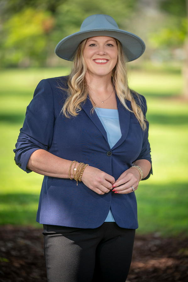 Young woman in blazer wearing a hat stands in front of a grassy area.