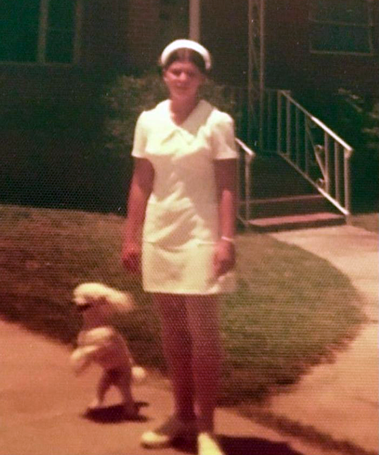A photo from the 1970s that shows a woman in a nurse's uniform. She is standing outside of a house.