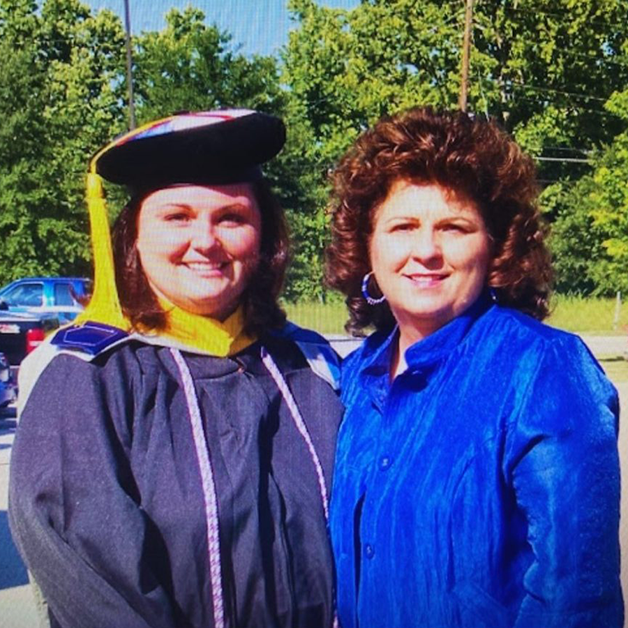 Two women stand outside and celebrate one graduating with her master's degree. The woman on the left is wearing full graduation regalia.