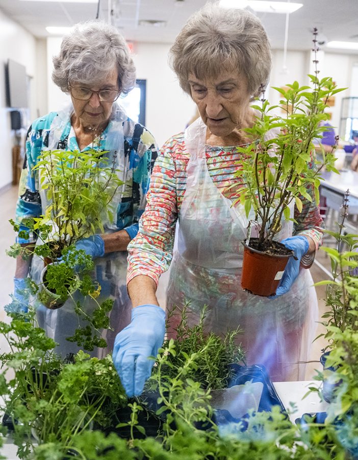 Two women wearing white aprons hold plants while looking at other plants