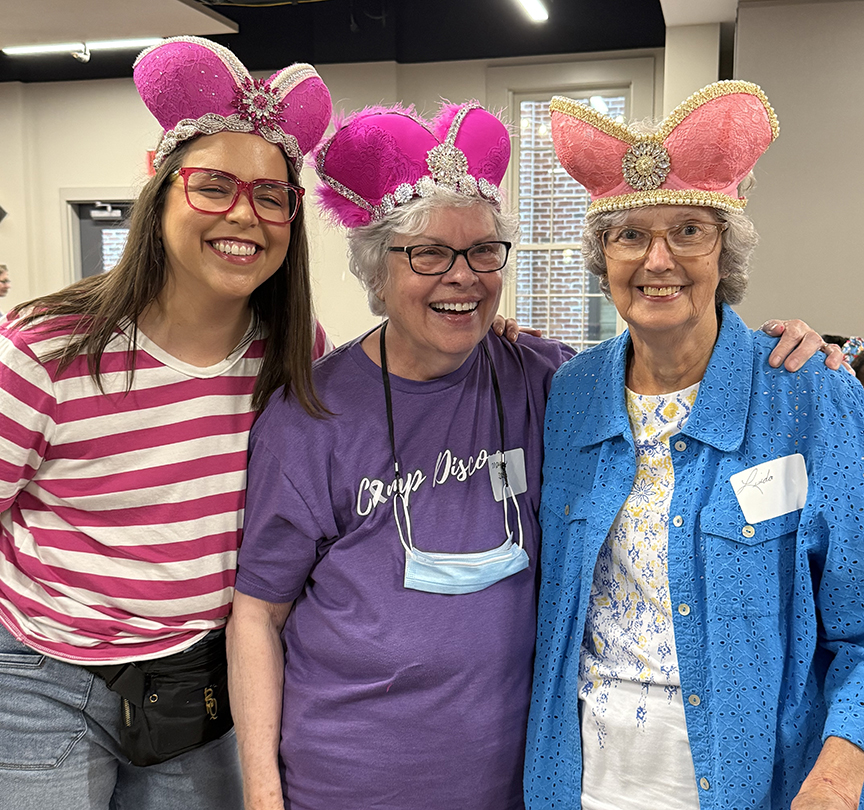 Three women wear crowns shaped like brassieres while laughing and smiling for the camera.