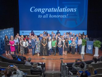 A large group of men and women stand on a stage with a large screen behind them. On the screen are the words "Congratulations to all honorees!"