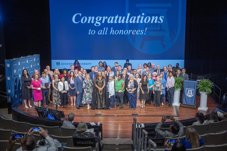 A large group of men and women stand on a stage with a large screen behind them. On the screen are the words "Congratulations to all honorees!"