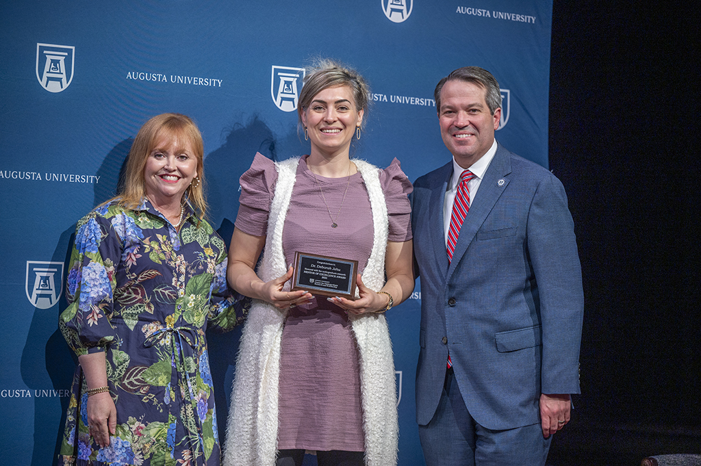 Two women and a man stand in front of an Augusta University backdrop. The woman in the middle is holding an award.