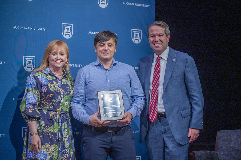 A woman and two men stand in front of an Augusta University backdrop. The man in the middle is holding an award.