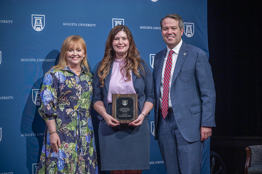 Two women and a man stand in front of an Augusta University backdrop. The woman in the middle is holding an award.