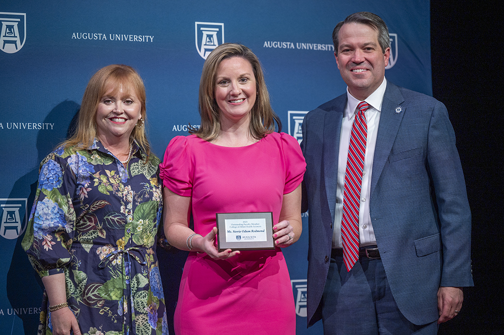 Two women and a man stand in front of an Augusta University backdrop. The woman in the middle is holding an award.