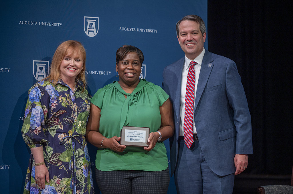 Two women and a man stand in front of an Augusta University backdrop. The woman in the middle is holding an award.