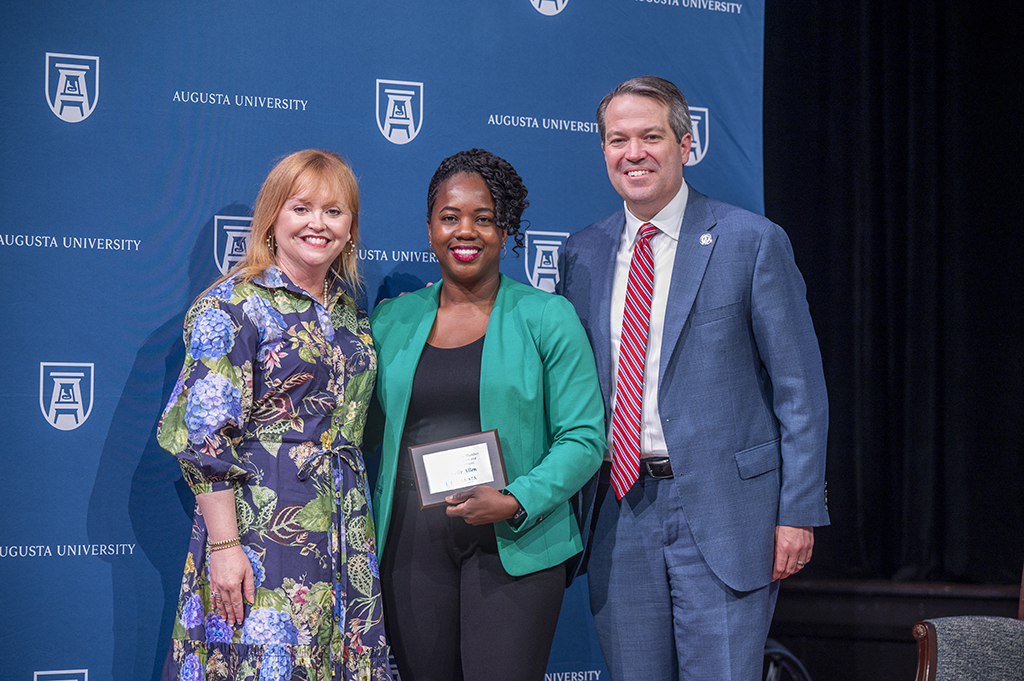Two women and a man stand in front of an Augusta University backdrop. The woman in the middle is holding an award.