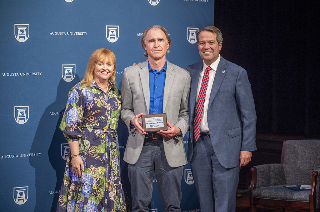 A woman and two men stand in front of an Augusta University backdrop. The man in the middle is holding an award.