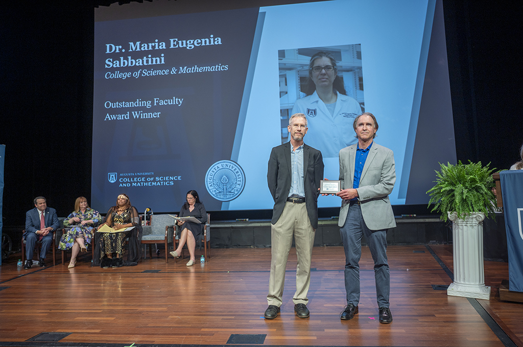 Two men stand on stage. The man on the right is holding an award. Behind them on a large screen is a photo of a woman on the right and text on the left that reads "Dr. Maria Eugenia Sabbatini, College of Science & Mathematics. Outstanding Faculty Award Winner." Under the text on the screen are three woman and a man sitting near the back of the stage.